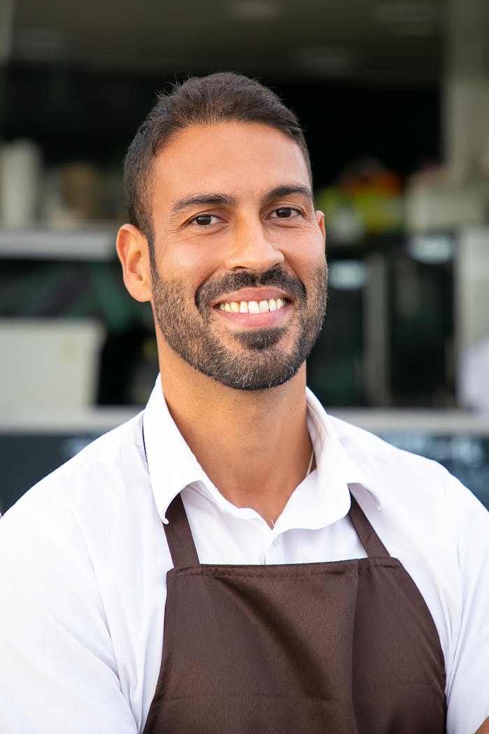 Cheerful ethnic waiter in apron standing against street cafe